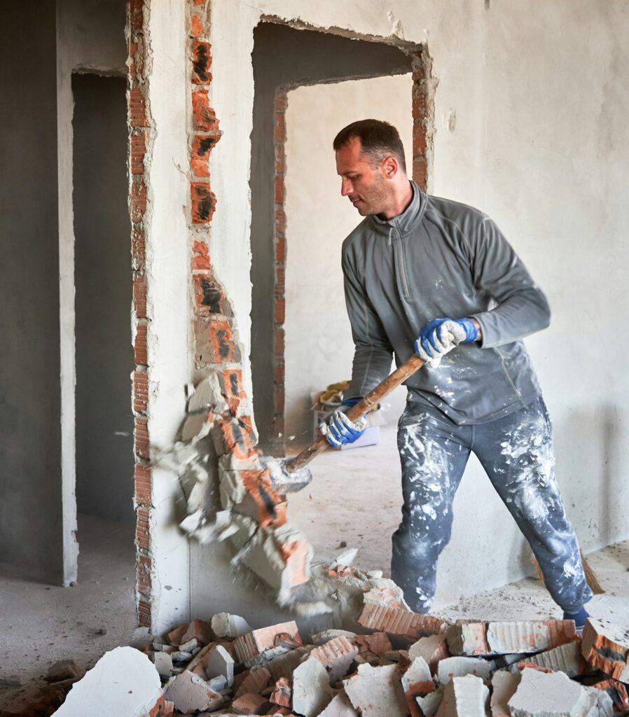 Male worker breaking wall with sledgehammer in apartment.